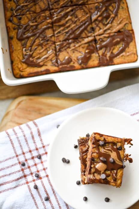 pumpkin blondies drizzles with melted chocolate and almond butter topped with chocolate chips on white plate beside a white pan of pumpkin blondies
