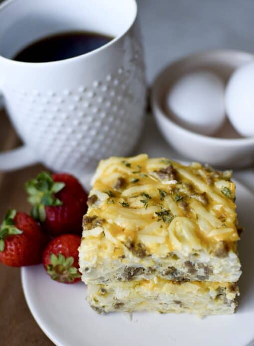 Sausage hashbrown casserole on white plate with strawberries, and cup of black coffee and eggs in the background 