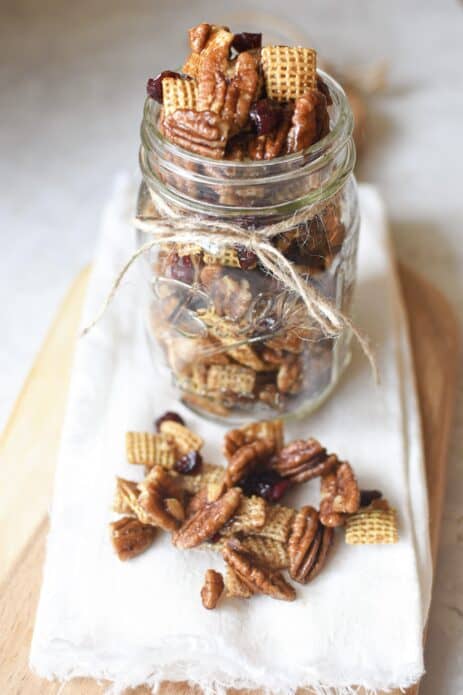clear mason jar with small twine ribbon tied around it, filled with pecan, chex mix and cranberry mixture sitting on white towel with pecan and chex mixture on towel