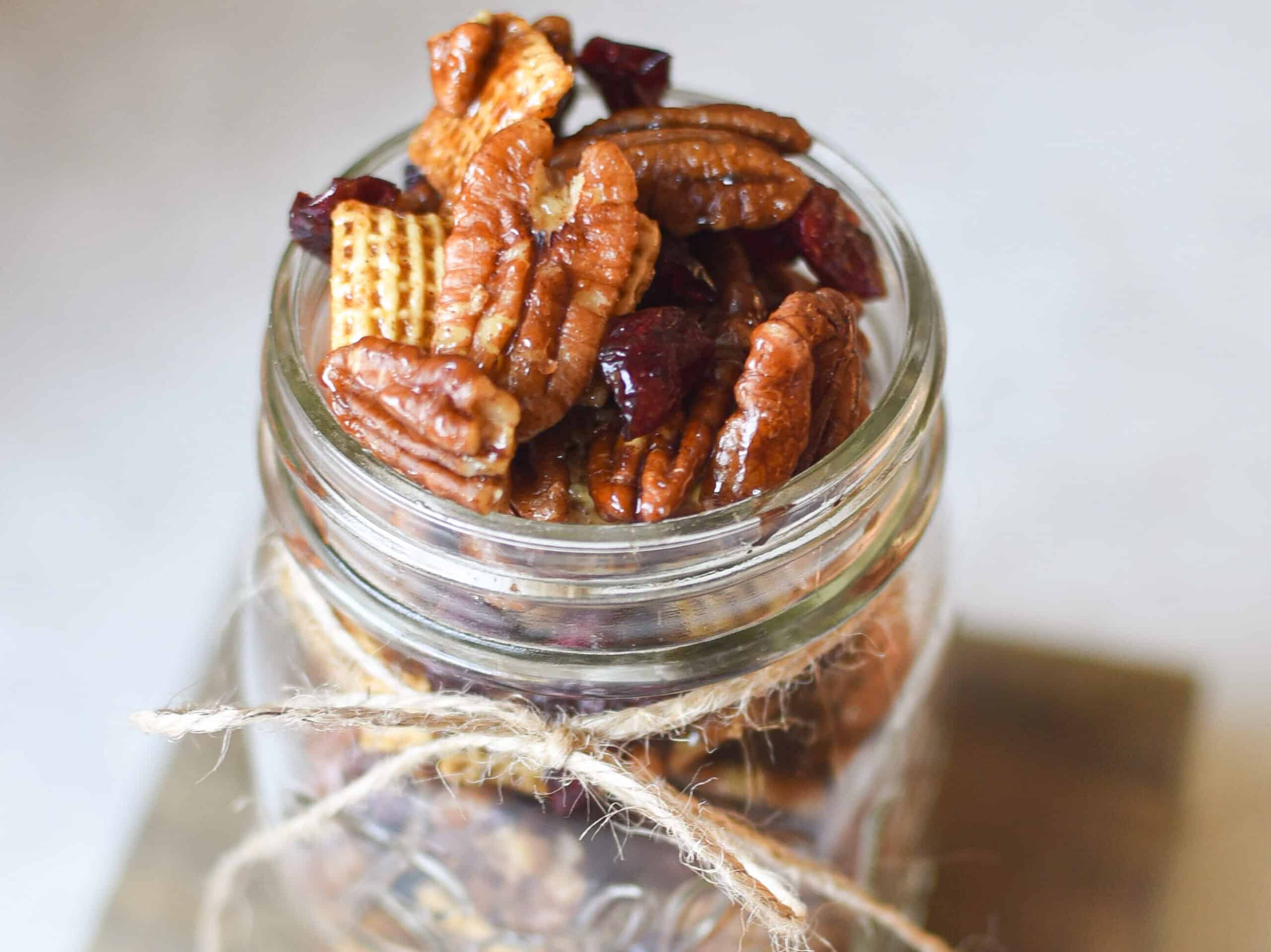 clear mason jar with small twine ribbon tied around it, filled with pecan, chex mix and cranberry mixture
