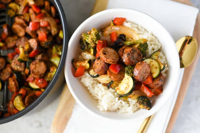 white bowl filled with chicken sausage and roasted vegetable medley over white rice with air fryer containing sausage and veggie mixture
