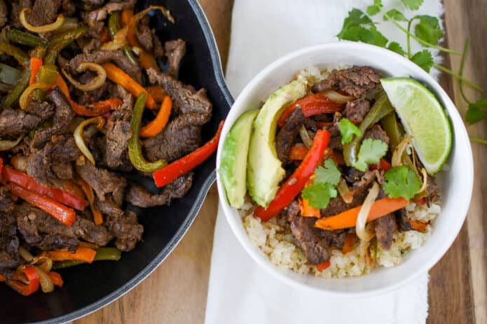 skillet filled with sliced steak, peppers and onions, beside a white bowl filled with cauliflower rice topped with sliced steak, peppers, onions, avocado, lime slice, and cilantro