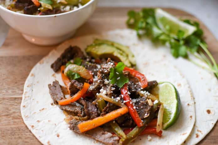 sliced steak, peppers and onions on flour tortillas with limes, avocado, and cilantro