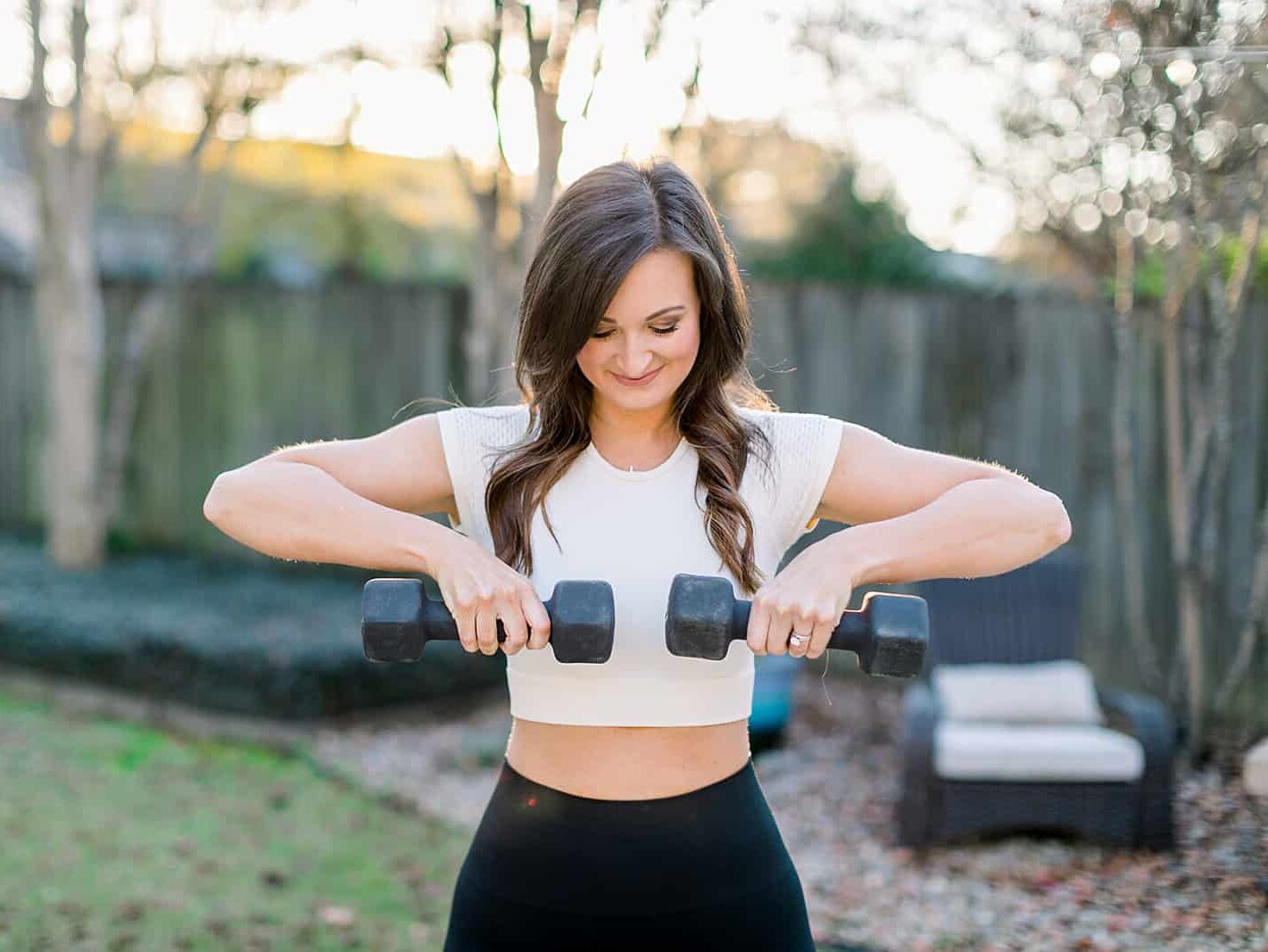 female with brown hair wearing white shirt lifting a dumbbell in each hand